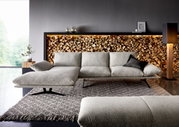Modernes hellgraues Stoffsofa mit linksseitiger Chaiselongue und schwarzen Metallfüßen auf Teppich vor dekorativer Holzscheitwand mit LED-Licht, Frontansicht in leicht schräger Perspektive.