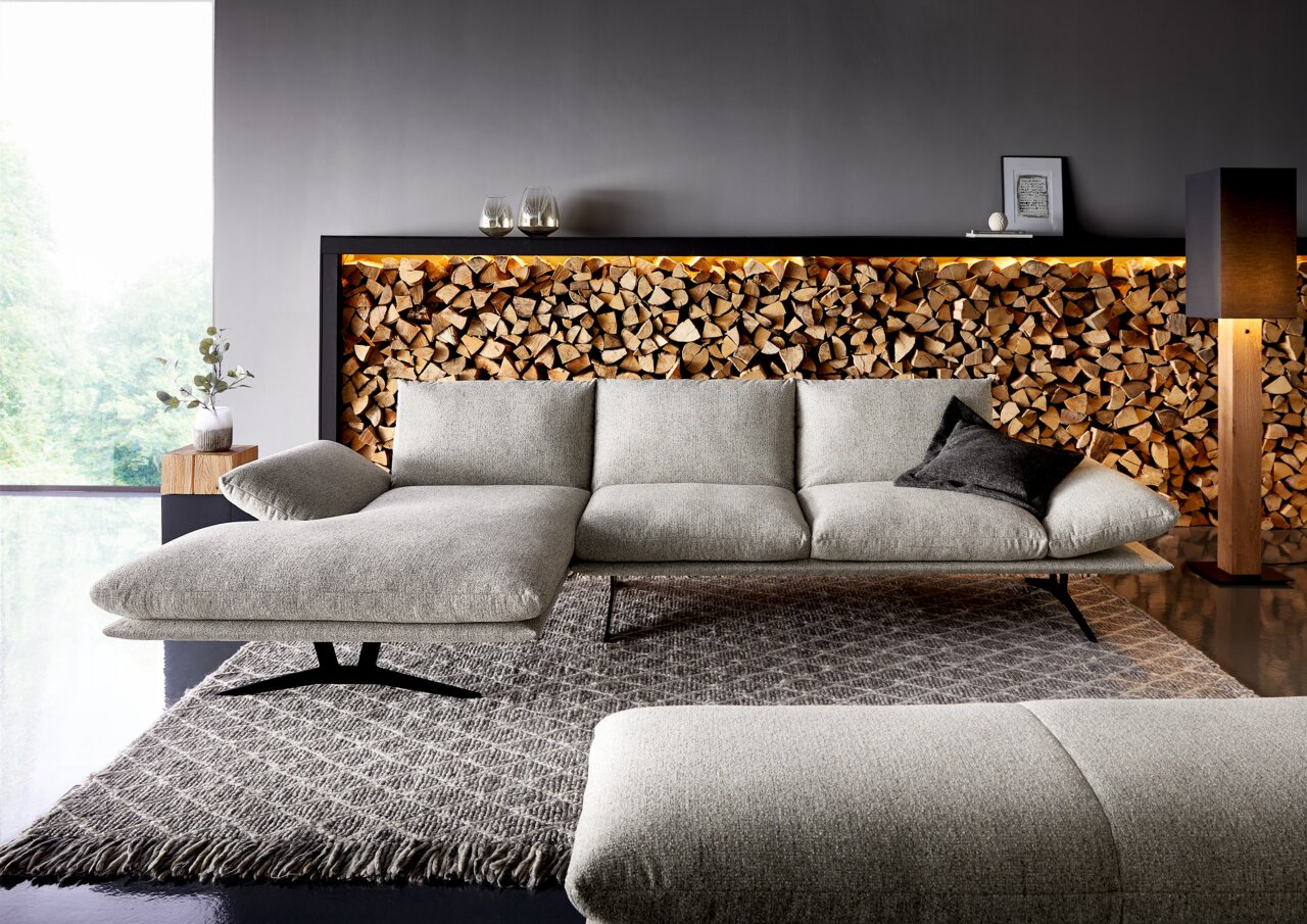 Modernes hellgraues Stoffsofa mit linksseitiger Chaiselongue und schwarzen Metallfüßen auf Teppich vor dekorativer Holzscheitwand mit LED-Licht, Frontansicht in leicht schräger Perspektive.