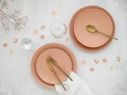 Draufsicht auf ein stilvolles Tischgedeck mit zwei roséfarbenen Tellern, goldfarbenem Besteck (Löffel, Messer, Gabel), zwei Wassergläsern, weißer Serviette und Konfetti auf hellem Hintergrund