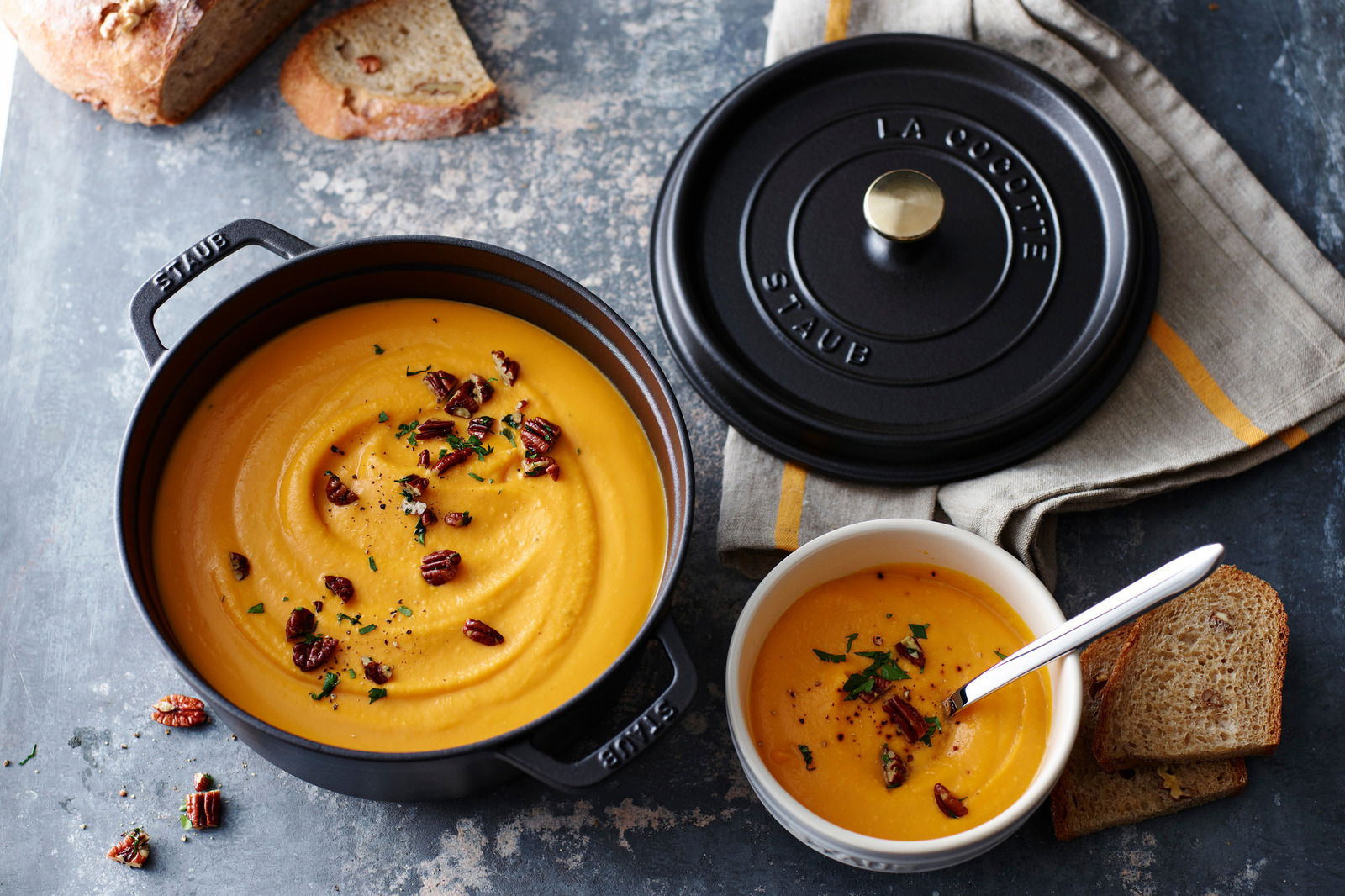 Runder Bräter Cocotte 24cm in Schwarz, gefüllt mit cremiger Suppe, von oben fotografiert. Daneben liegt der Deckel auf einem Tuch.