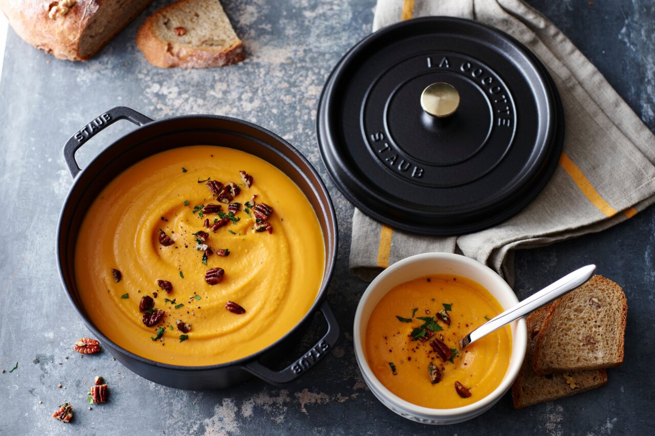 Runder Bräter Cocotte 24cm in Schwarz, gefüllt mit cremiger Suppe, von oben fotografiert. Daneben liegt der Deckel auf einem Tuch.
