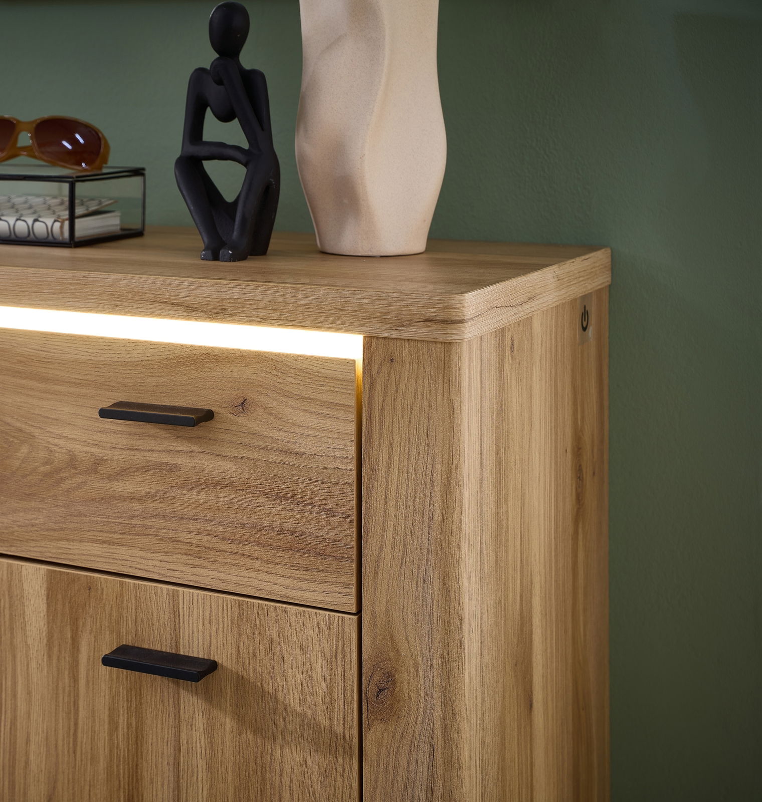 Nahaufnahme eines modernen Holzschranks mit LED-Beleuchtung, schwarzem Griff und dekorativen Objekten auf der Oberseite, seitliche Perspektive.