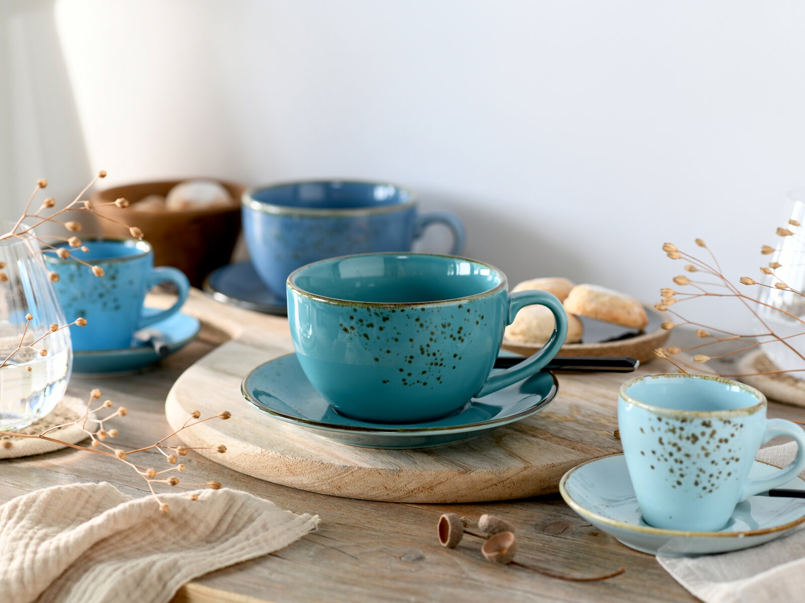 Türkise, gesprenkelte Keramik-Teetasse mit Untertasse auf Holzbrett, seitliche Nahaufnahme auf Tischgedeck mit weiteren Tassen im Hintergrund (Perspektive: leicht seitlich von vorn, Nahaufnahme).