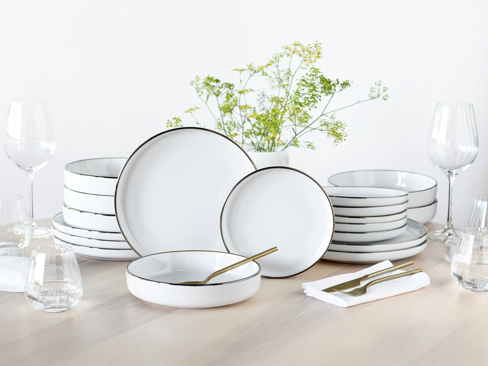 Elegantes weißes Geschirrset aus Porzellan mit feinem goldfarbenem Rand, bestehend aus Tellern und Schalen, auf gedecktem Holztisch mit Gläsern und goldenem Besteck, Perspektive: frontale leicht erhöhte Ansicht.