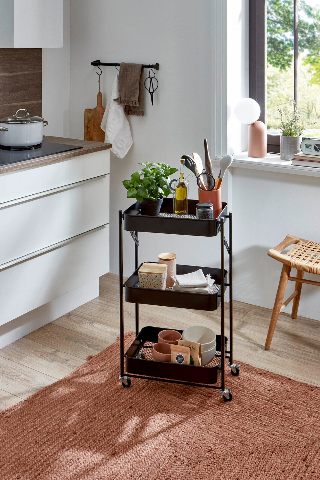 Schwarzer, dreistöckiger Küchen-Servierwagen aus Metall mit Rollen, in einer hellen Küche mit Pflanzen und Küchenutensilien bestückt, fotografiert in leichter Seitenansicht auf Augenhöhe.