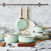Set aus mintgrünen Bratpfannen und Töpfen mit goldenen Griffen, präsentiert auf einer Küchenarbeitsplatte vor einer weißen Kachelwand, Perspektive von vorne.