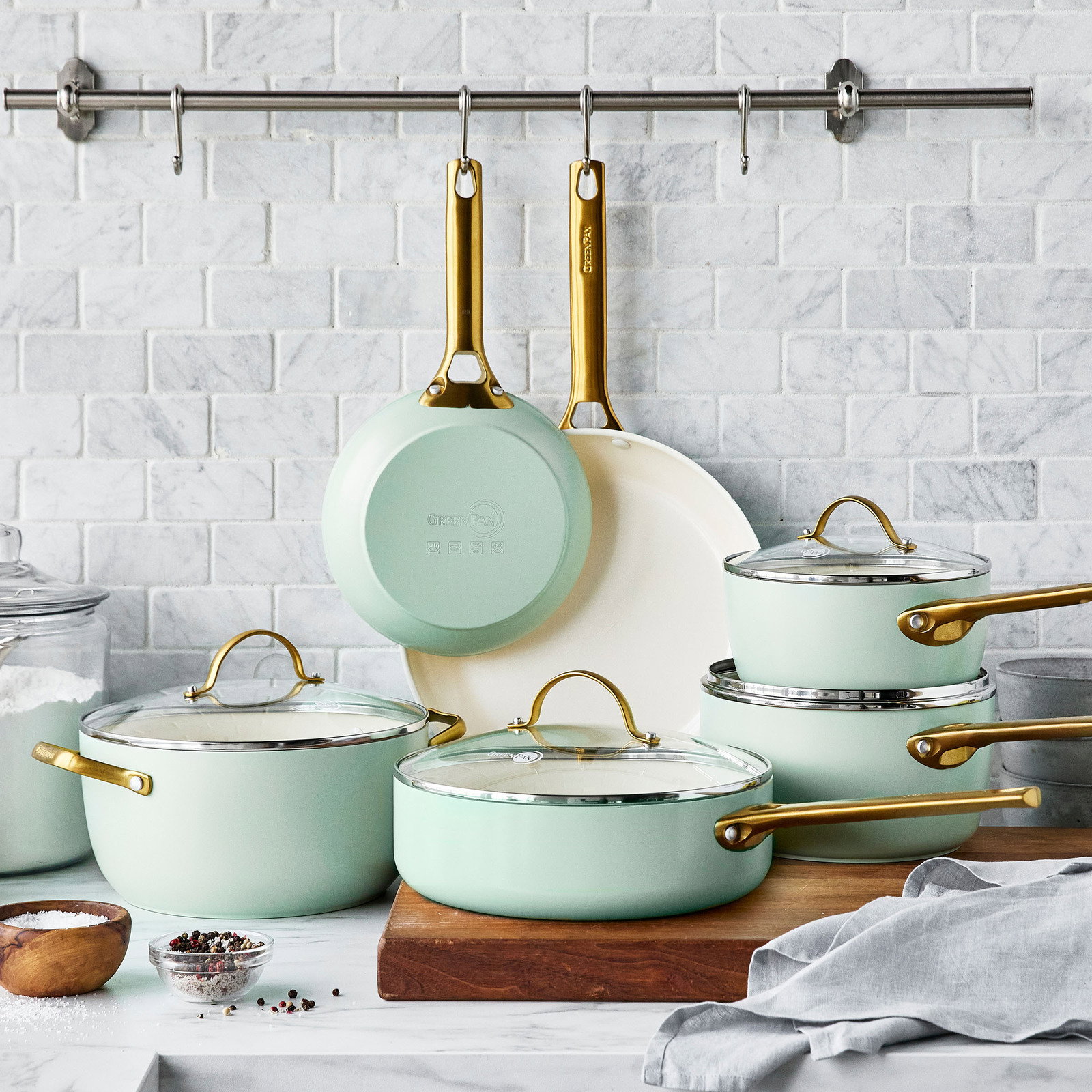 Set aus mintgrünen Bratpfannen und Töpfen mit goldenen Griffen, präsentiert auf einer Küchenarbeitsplatte vor einer weißen Kachelwand, Perspektive von vorne.