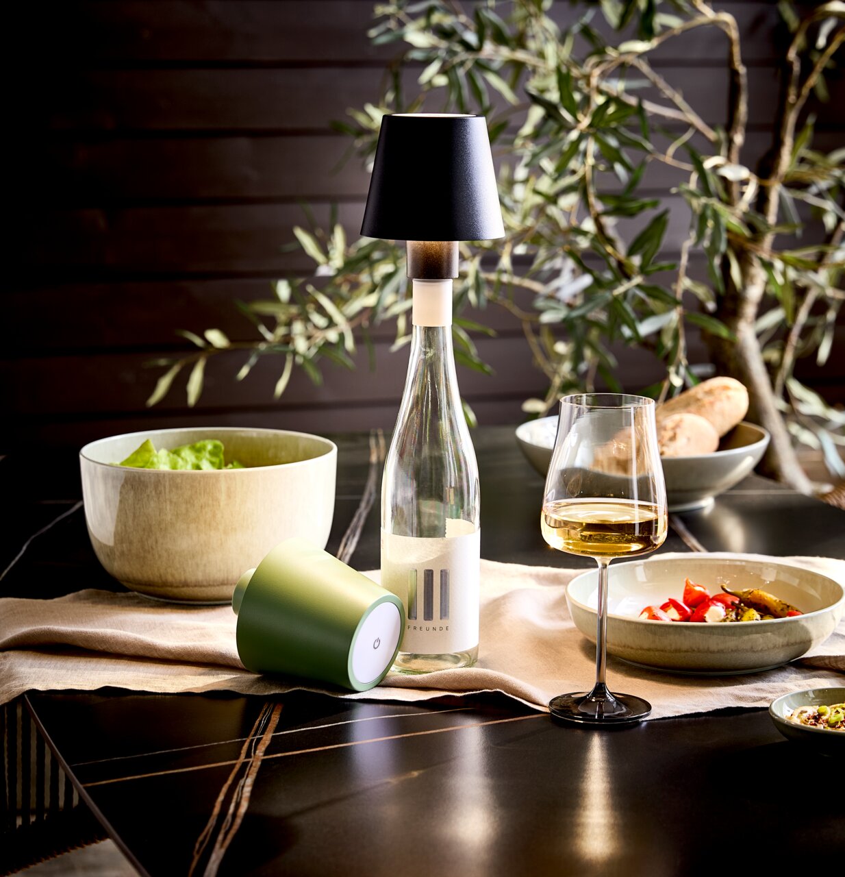Akku-Tischleuchte Novel auf einem gedeckten Tisch, seitliche Perspektive, mit Weinflasche und Glas.