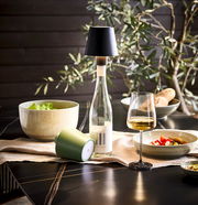 Akku-Tischleuchte Novel auf einem gedeckten Tisch, seitliche Perspektive, mit Weinflasche und Glas.