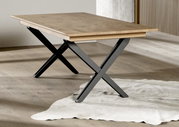 Moderne eettafel met een lichtgekleurd houten blad en zwarte X-vormige metalen poten in een minimalistische ruimte, schuin van voren gefotografeerd vanuit een iets hoger standpunt.