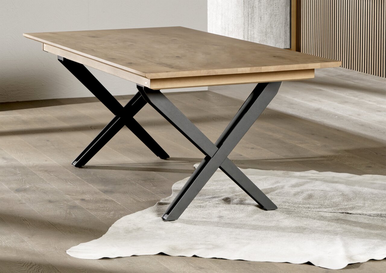Moderne eettafel met een lichtgekleurd houten blad en zwarte X-vormige metalen poten in een minimalistische ruimte, schuin van voren gefotografeerd vanuit een iets hoger standpunt.