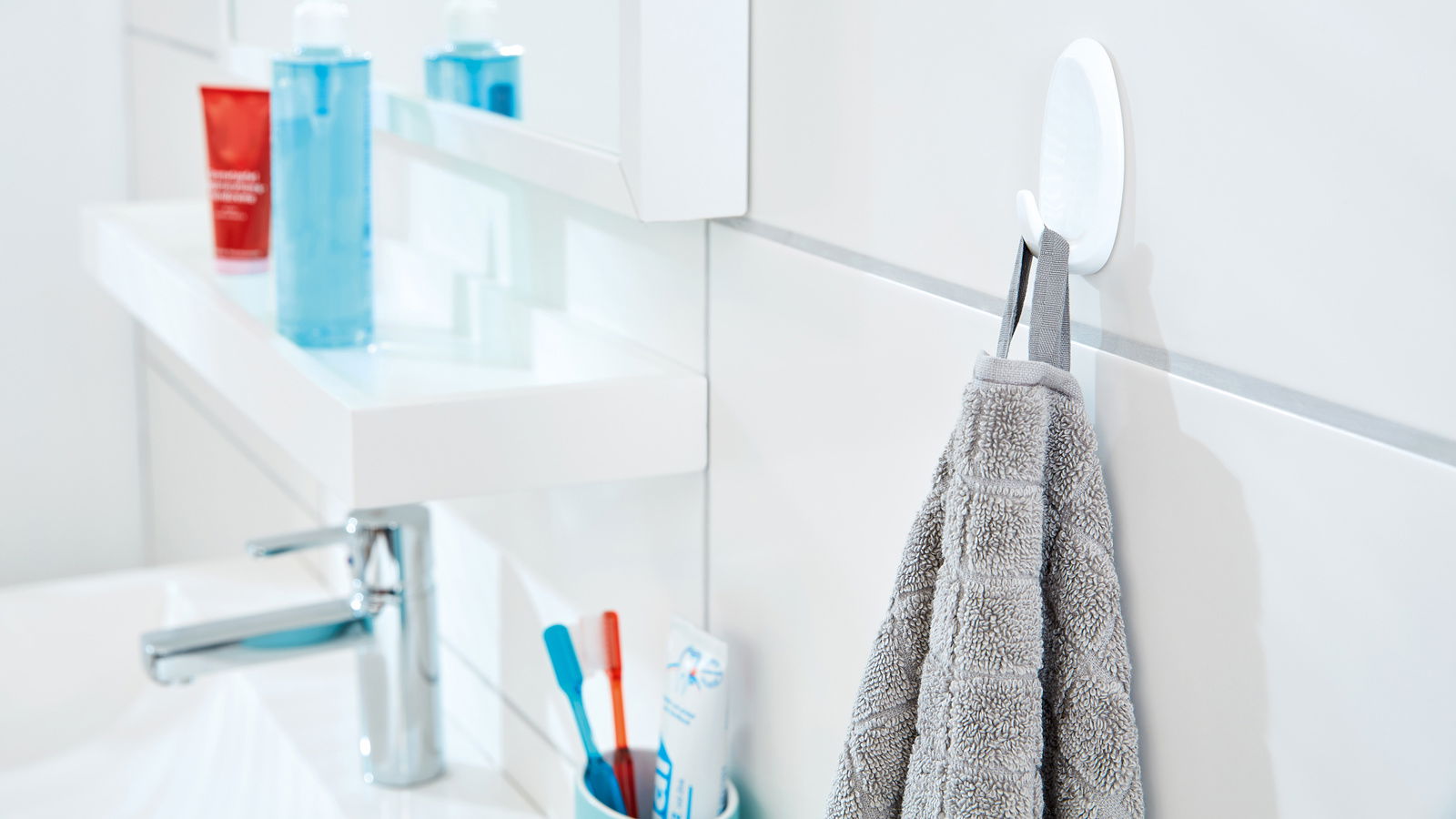 Ein weißer tesa Powerstrips Haken Large an einer Badezimmerwand, der ein graues Handtuch hält. Perspektive von der Seite.