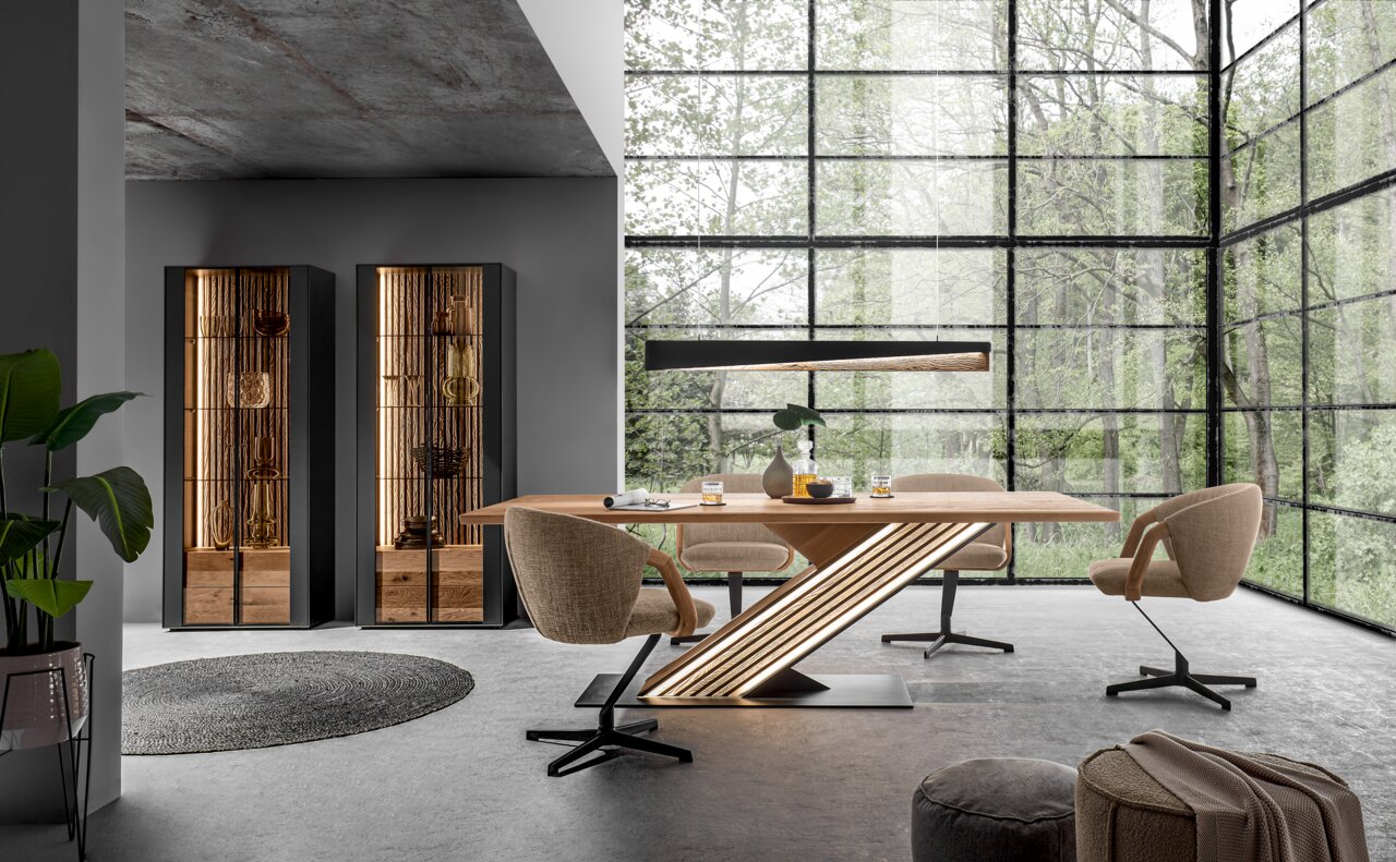 Johann Jakob Esstisch LUCIANO Moderne Esszimmer-Szene mit großem Holztisch und LED-Beleuchtung, umgeben von drehbaren Stühlen und hohen Vitrinen, mit Blick auf eine grüne Außenlandschaft durch große Fenster.