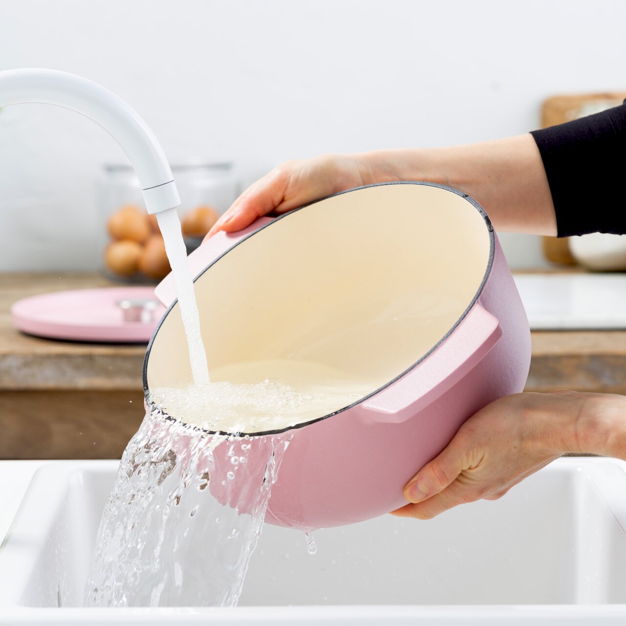 Rosa Gusseisen Bräter, rund, 26 cm, wird in einem Waschbecken unter fließendem Wasser gereinigt, seitliche Perspektive.