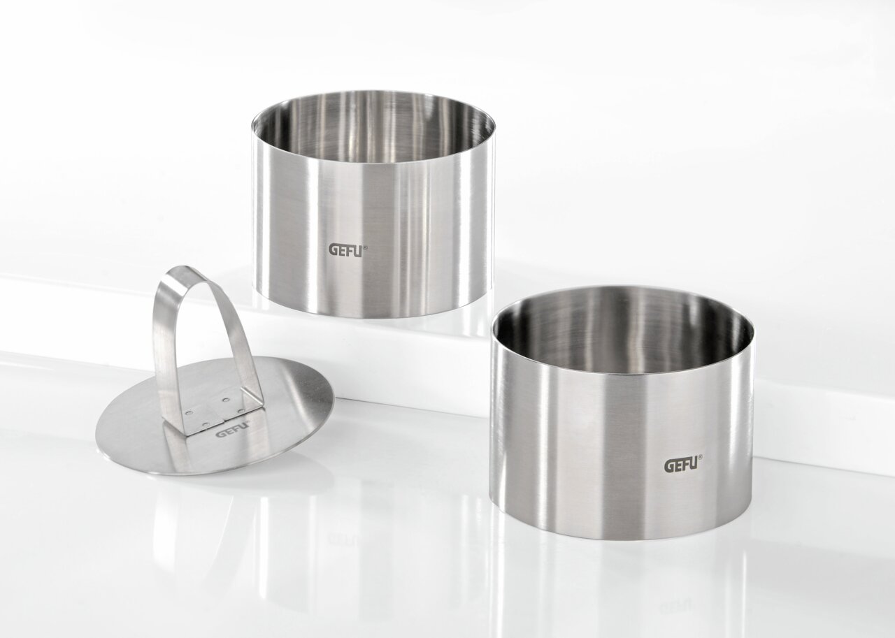 Zwei Edelstahlringe und ein Edelstahlpresser, frontal und leicht von oben betrachtet, ideal für Desserts oder Vorspeisen.