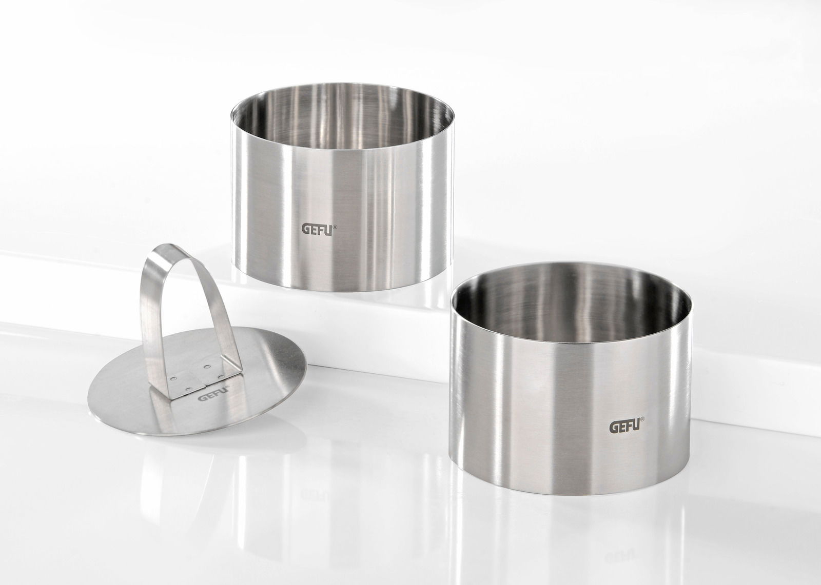 Zwei Edelstahlringe und ein Edelstahlpresser, frontal und leicht von oben betrachtet, ideal für Desserts oder Vorspeisen.