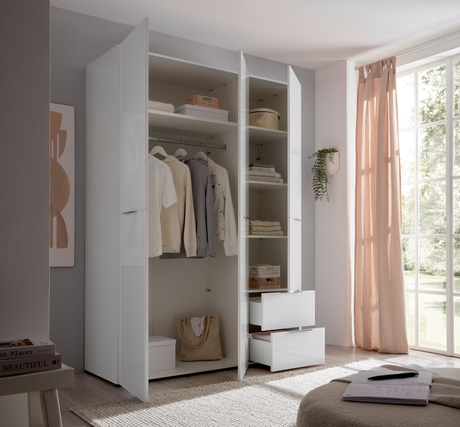 Offener weißer Kleiderschrank mit glänzenden Fronten, Kleiderstange, Regalböden und zwei Auszügen, seitliche Frontansicht im hellen Schlafzimmer neben großem Fenster mit rosa Vorhang