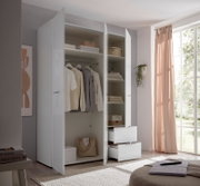 Offener weißer Kleiderschrank mit glänzenden Fronten, Kleiderstange, Regalböden und zwei Auszügen, seitliche Frontansicht im hellen Schlafzimmer neben großem Fenster mit rosa Vorhang