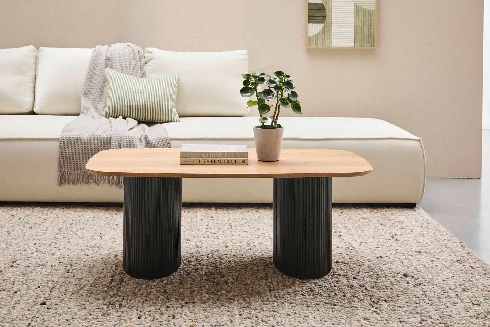 Stylife Couchtisch GROOVE WOOD Moderner Couchtisch mit ovaler Holzplatte und zwei schwarzen, geriffelten Säulenbeinen, frontal vor einem cremefarbenen Sofa mit Kissen und Decke, auf einem gemusterten Teppich.