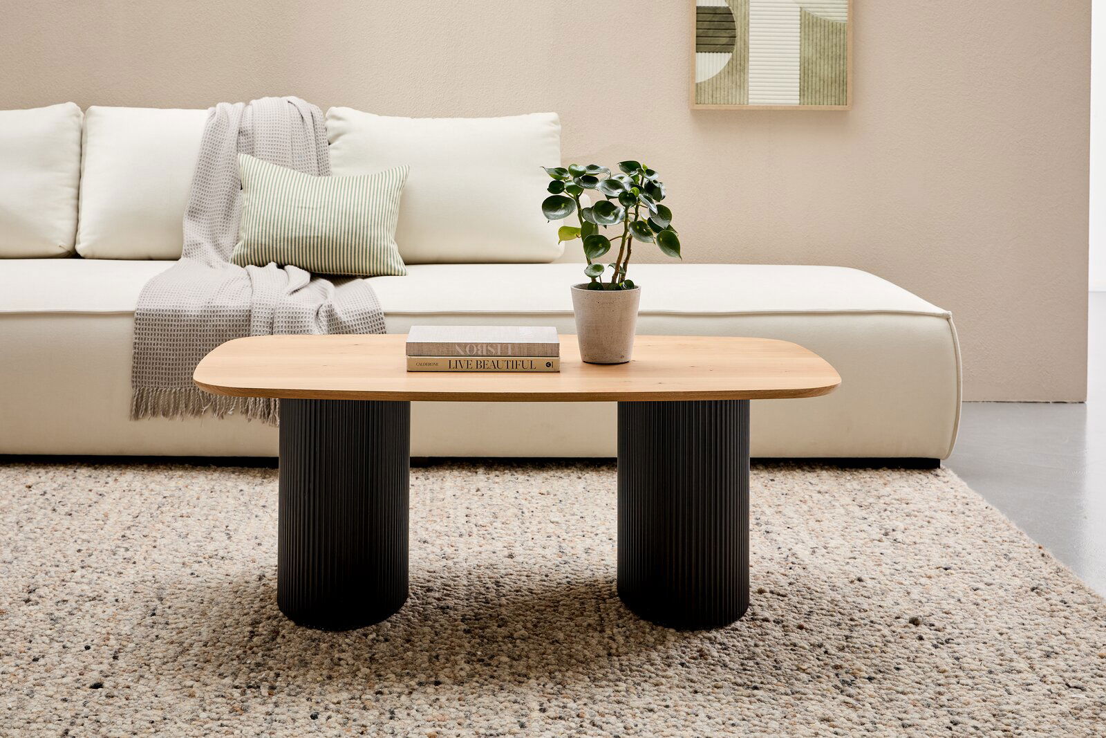 Stylife Couchtisch GROOVE WOOD Moderner Couchtisch mit ovaler Holzplatte und zwei schwarzen, geriffelten Säulenbeinen, frontal vor einem cremefarbenen Sofa mit Kissen und Decke, auf einem gemusterten Teppich.