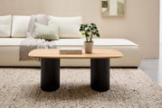 Moderner Couchtisch mit ovaler Holzplatte und zwei schwarzen, geriffelten Säulenbeinen, frontal vor einem cremefarbenen Sofa mit Kissen und Decke, auf einem gemusterten Teppich.