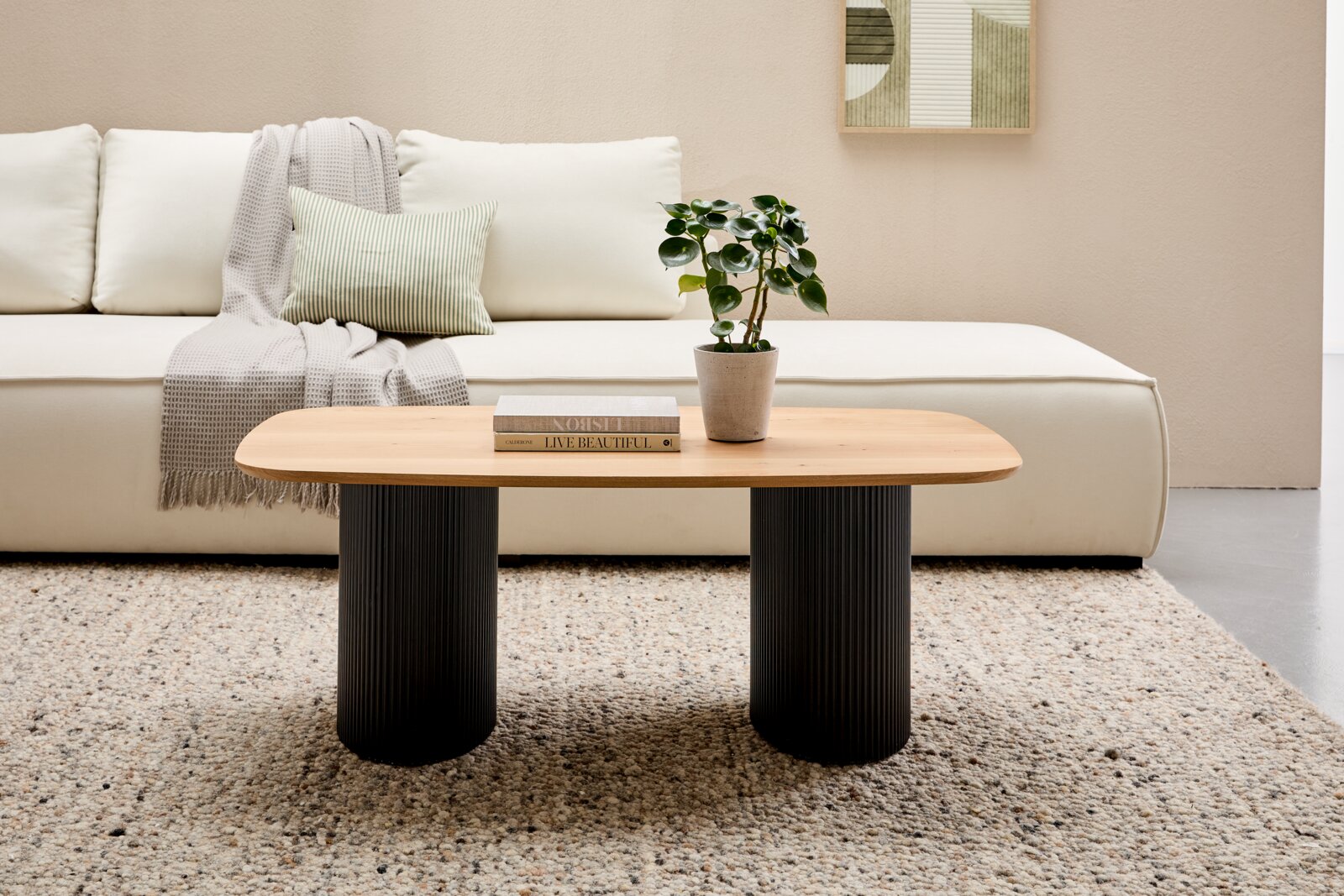 Stylife Salontafel GROOVE WOOD Moderne salontafel met een ovaal houten blad en twee zwarte, geribbelde kolomvormige poten, gezien van voren voor een crèmekleurige bank met kussens en een deken, op een tapijt met patroon.