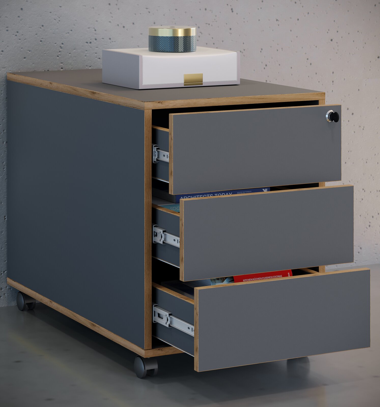 Moderner Rollcontainer mit drei halb geöffneten Schubladen, anthrazitgraue Fronten und Korpus mit Holzrahmen, auf der Deckplatte eine weiße Box und ein kleiner runder Lautsprecher Ansicht schräg von vorne links.