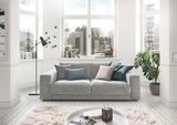 Graues 2-Sitzer-Sofa in einem hellen Wohnzimmer mit großen Fenstern, dekoriert mit bunten Kissen. Perspektive von vorne.