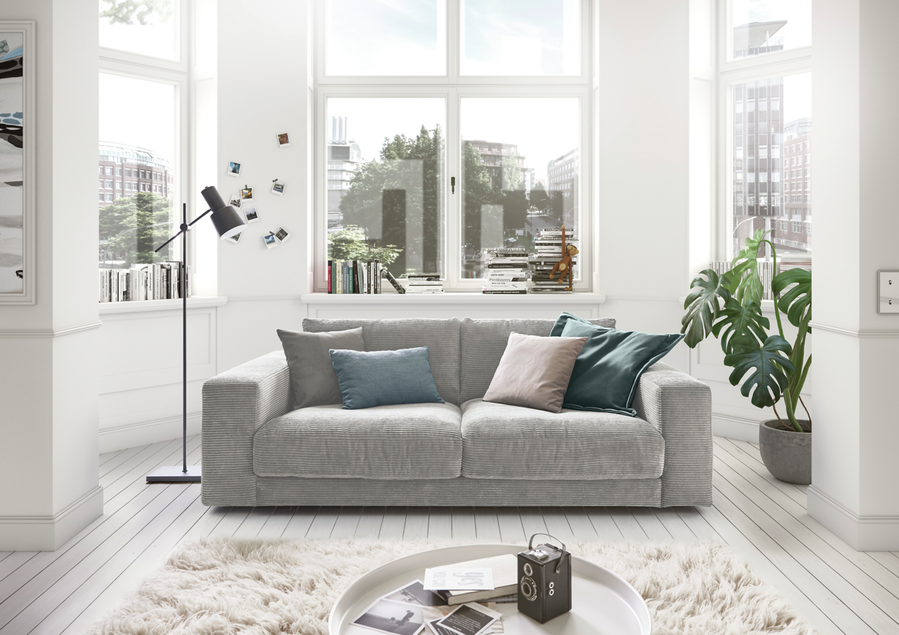 Lounge Handmade Sofa 2-Sitzer LH-SAMBIA Graues 2-Sitzer-Sofa in einem hellen Wohnzimmer mit großen Fenstern, dekoriert mit bunten Kissen. Perspektive von vorne.