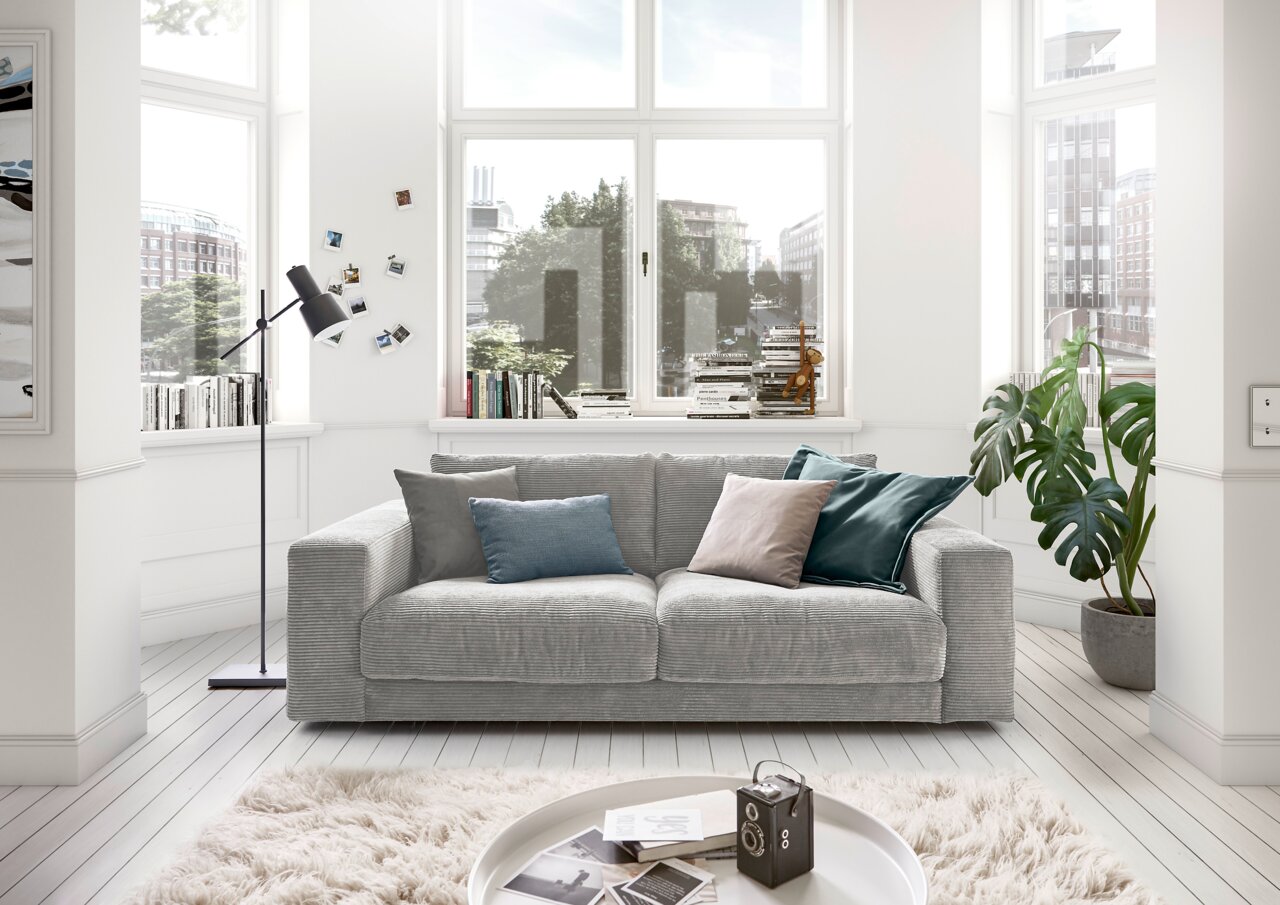 Graues 2-Sitzer-Sofa in einem hellen Wohnzimmer mit großen Fenstern, dekoriert mit bunten Kissen. Perspektive von vorne.