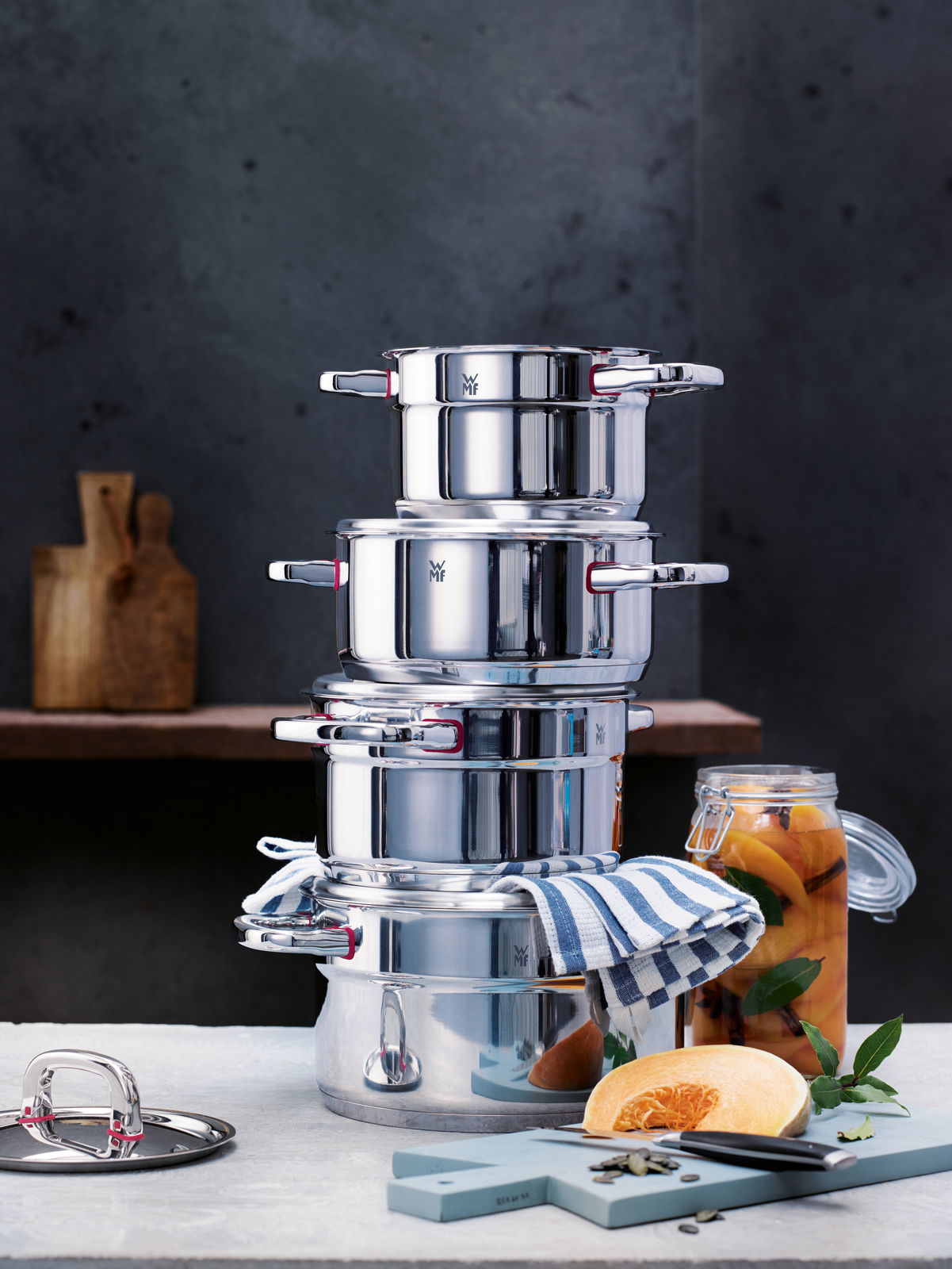 Stapel von glänzenden Edelstahl-Töpfen aus dem Topfset Premium One 5-tlg., seitliche Perspektive, mit einem gestreiften Küchentuch und einem Glas mit eingelegtem Gemüse im Hintergrund.