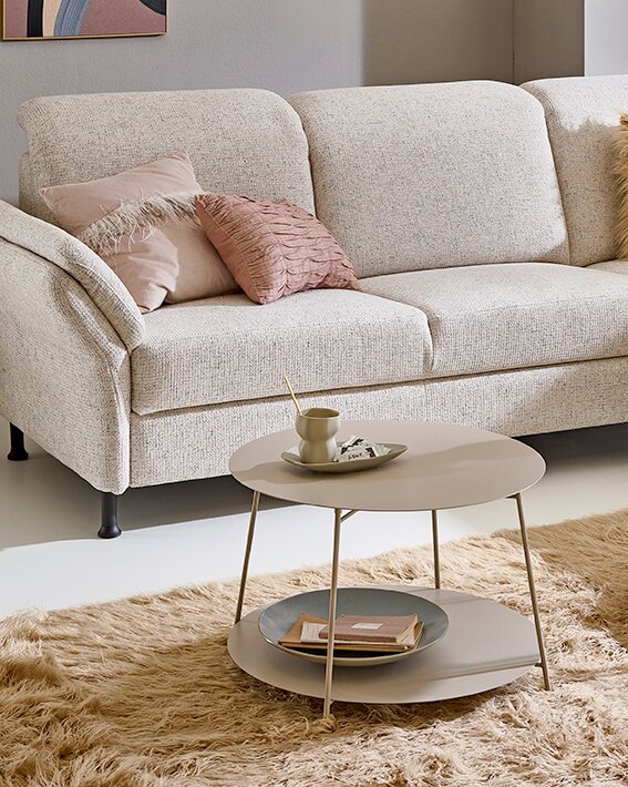 Beiger runder Couchtisch mit zwei Ebenen und schlanken Metallbeinen auf flauschigem Teppich vor hellem Sofa, fotografiert aus leicht erhöhter Frontperspektive.