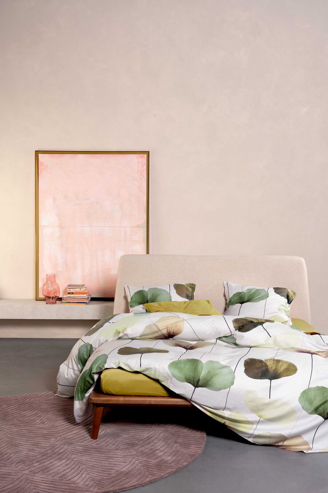 Essenza Home Bettwäsche KATI Modernes Schlafzimmer mit Bett in beiger Polsteroptik und Bettwäsche mit großem Blattmuster in Grün- und Beigetönen, aufgenommen in frontaler Perspektive leicht von links; im Hintergrund ein gerahmtes Bild, Bücherstapel und rosa Glasvase auf einer Konsole