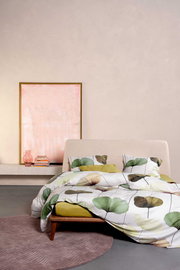 Modernes Schlafzimmer mit Bett in beiger Polsteroptik und Bettwäsche mit großem Blattmuster in Grün- und Beigetönen, aufgenommen in frontaler Perspektive leicht von links; im Hintergrund ein gerahmtes Bild, Bücherstapel und rosa Glasvase auf einer Konsole