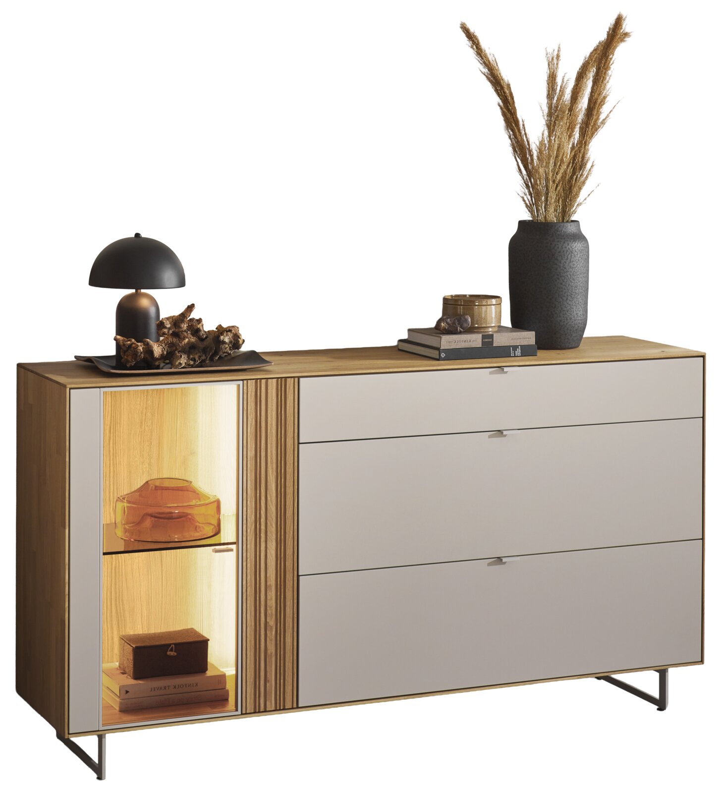 Modernes Sideboard aus Holz mit hellen Schubladenfronten und links beleuchteter Vitrine, dekoriert mit Lampe, Büchern und Vase mit Trockenblumen, frontale Perspektive