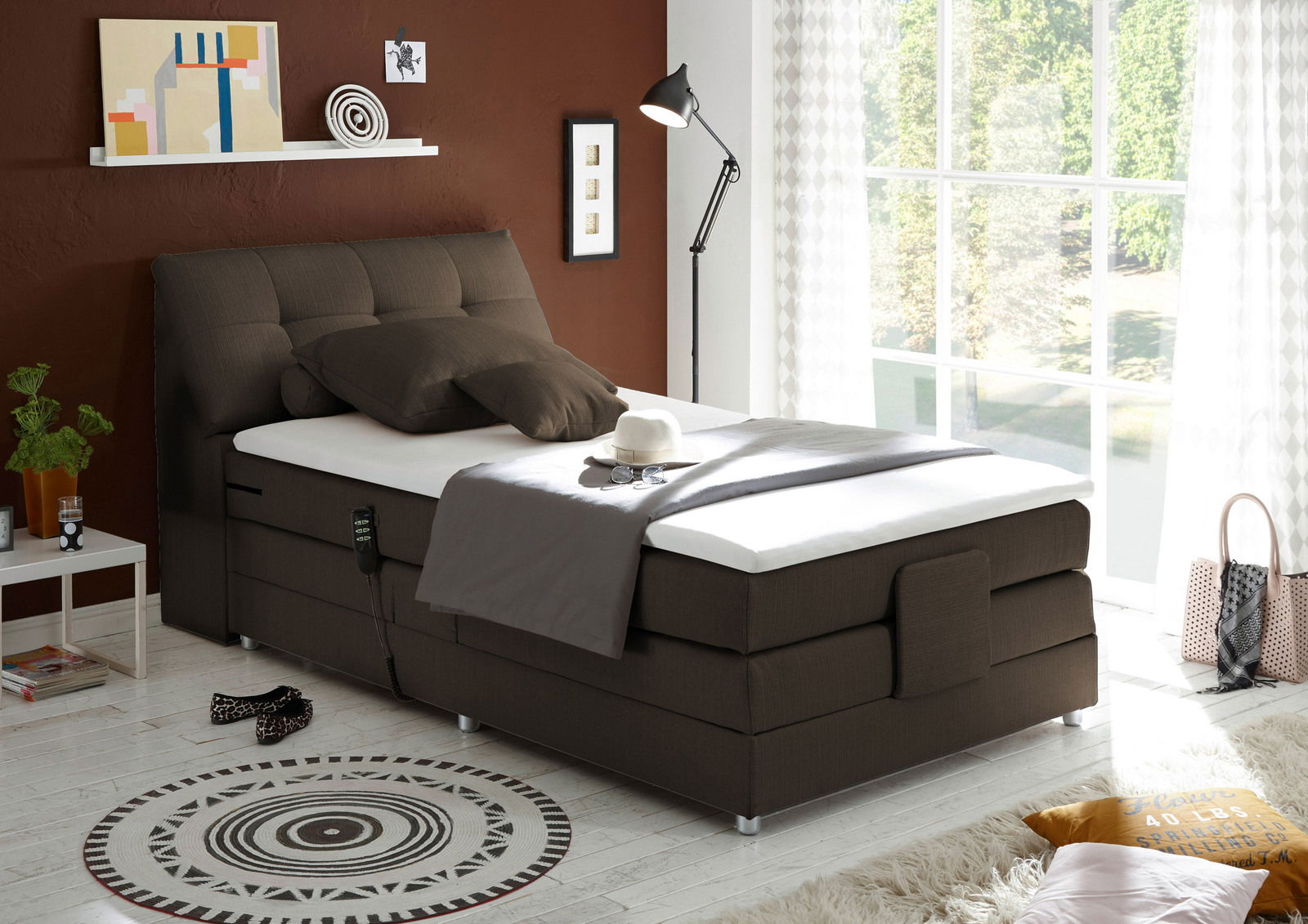 Boxspringbett in einem modernen Schlafzimmer, seitliche Perspektive, mit braunem Kopfteil und weißer Matratze, dekoriert mit Kissen und Decke, neben einem Fenster mit Blick ins Grüne.