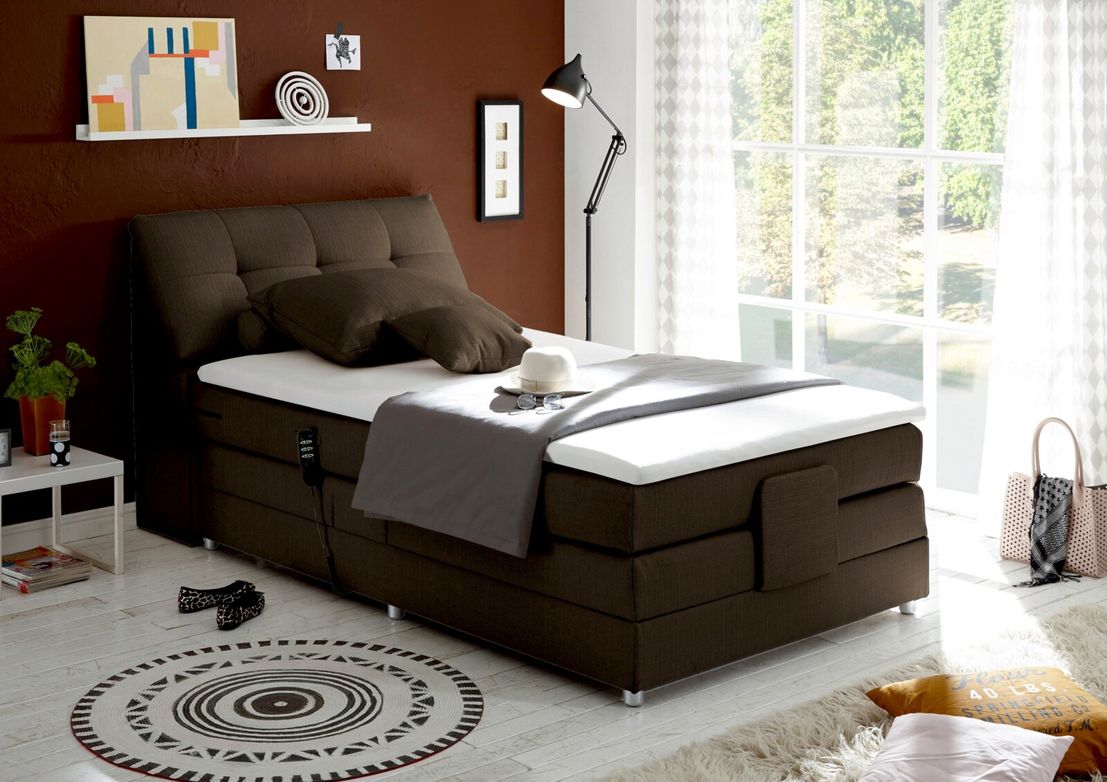 Boxspringbett in einem modernen Schlafzimmer, seitliche Perspektive, mit braunem Kopfteil und weißer Matratze, dekoriert mit Kissen und Decke, neben einem Fenster mit Blick ins Grüne.