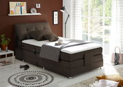 Boxspringbett in einem modernen Schlafzimmer, seitliche Perspektive, mit braunem Kopfteil und weißer Matratze, dekoriert mit Kissen und Decke, neben einem Fenster mit Blick ins Grüne.