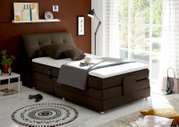Boxspringbett in einem modernen Schlafzimmer, seitliche Perspektive, mit braunem Kopfteil und weißer Matratze, dekoriert mit Kissen und Decke, neben einem Fenster mit Blick ins Grüne.