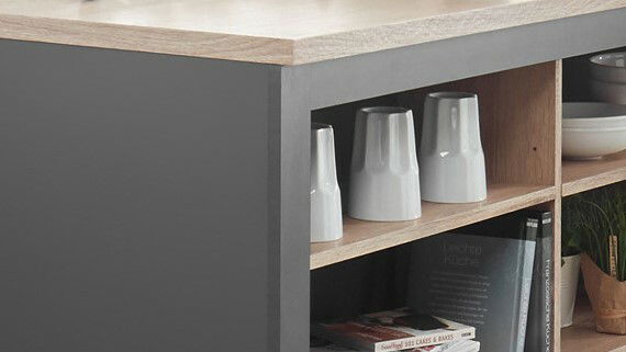 Kücheninsel mit grauer Seitenverkleidung und Holzregalen, gefüllt mit weißen Bechern und Büchern, aus seitlicher Perspektive.