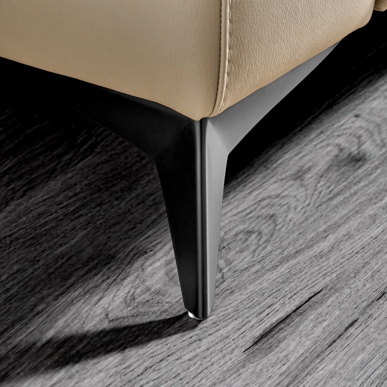 Nahaufnahme eines modernen Möbelfußes aus schwarzem Metall an einem beigen Ledermöbelstück, perspektivisch von unten aufgenommen.