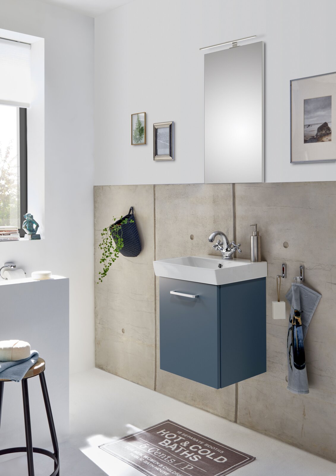 Modernes Badezimmer mit wandhängendem Waschtischunterschrank in Dunkelblau, weißem Waschbecken, verchromter Armatur und Spiegel mit Aufsatzleuchte, fotografiert in leichter Seitenansicht von vorn.