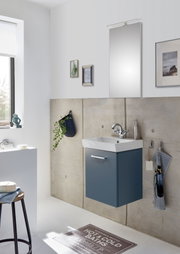 Modernes Badezimmer mit wandhängendem Waschtischunterschrank in Dunkelblau, weißem Waschbecken, verchromter Armatur und Spiegel mit Aufsatzleuchte, fotografiert in leichter Seitenansicht von vorn.