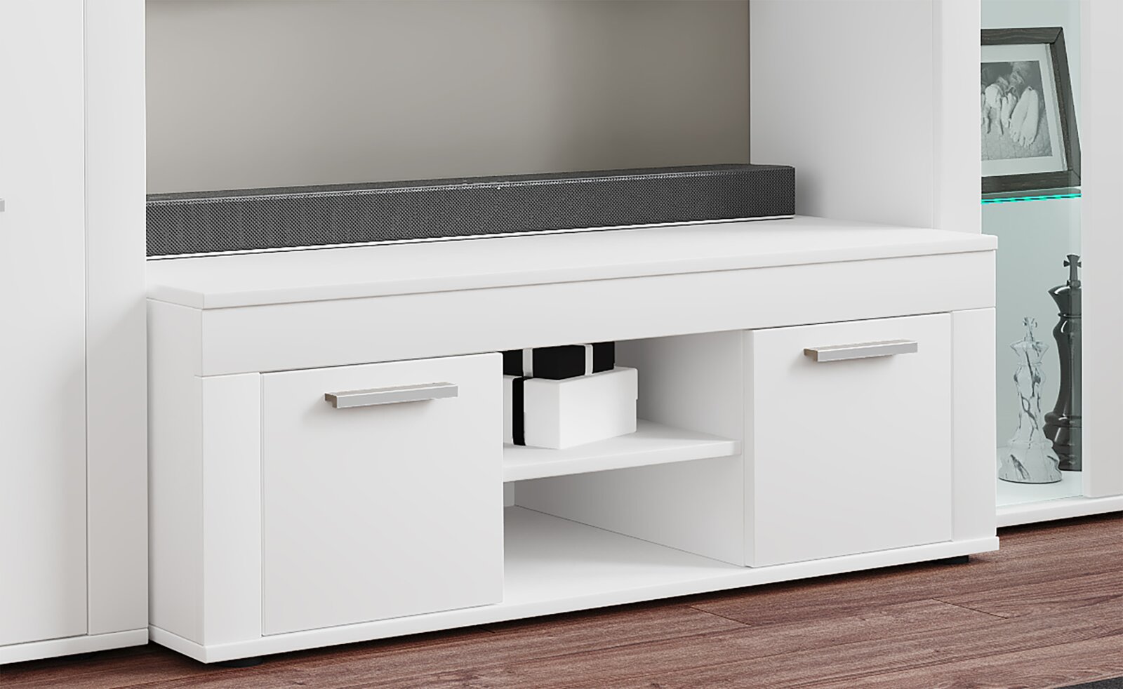 Weißes TV-Lowboard mit zwei Schranktüren, offenem Mittelregal und silbernen Griffen, mit schwarzer Soundbar auf der Ablage, Ansicht von vorne in leichter Schrägperspektive.