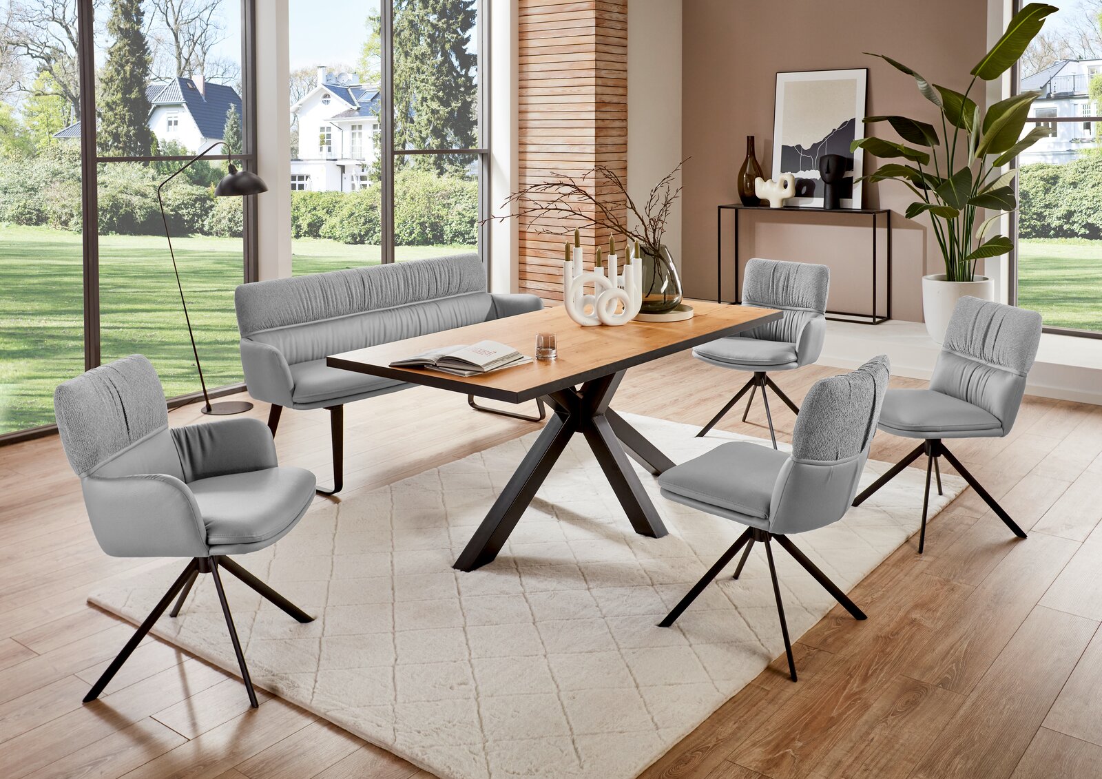 Moderner Esstisch mit Holzplatte und schwarzen Metallbeinen, umgeben von grauen Stühlen und einer Bank, in einem hellen Raum mit großen Fenstern und Blick auf den Garten, aus seitlicher Perspektive.