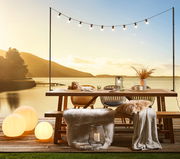 Gartentisch auf einer Terrasse mit Blick auf einen See bei Sonnenuntergang, dekoriert mit Lichterkette, Kissen und Decken.