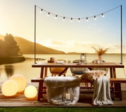 Gartentisch auf einer Terrasse mit Blick auf einen See bei Sonnenuntergang, dekoriert mit Lichterkette, Kissen und Decken.