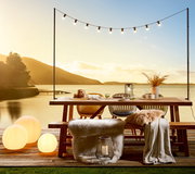 Gartentisch auf einer Terrasse mit Blick auf einen See bei Sonnenuntergang, dekoriert mit Lichterkette, Kissen und Decken.