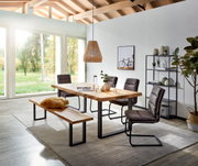 Moderne eetkamerset met een massief houten eettafel en een bijpassende houten bank op een zwart metalen onderstel, aangevuld met vier grijze zweefstoelen, gefotografeerd vanuit een licht schuin perspectief van voren in een lichte ruimte met grote ramen