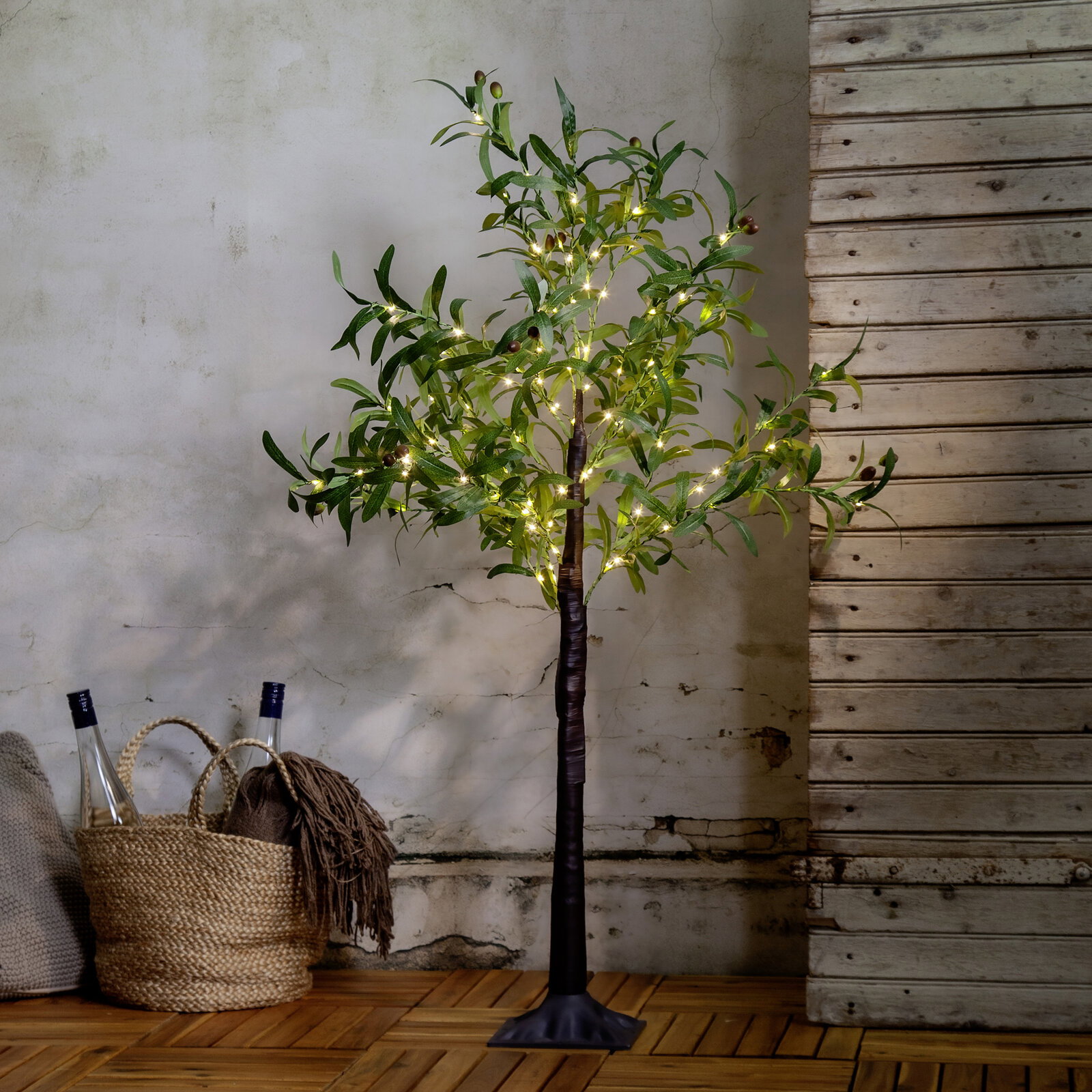 Dekorativer künstlicher Olivenbaum mit warmweißer LED-Lichterkette und schwarzem Standfuß, frontal in Innenraumkulisse vor rustikaler Wand und Holzboden.